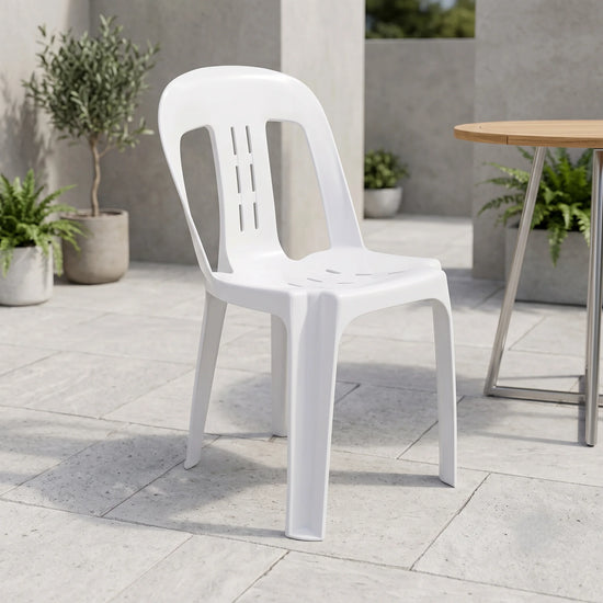 A white plastic outdoor dining chair with a slatted back and seat, shown on a light stone patio next to a wooden table.